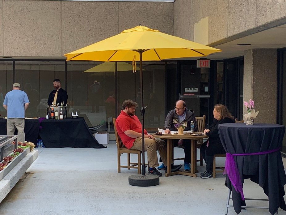 Three people seated at a table under an umbrella, with a drink table and other decorations nearby.