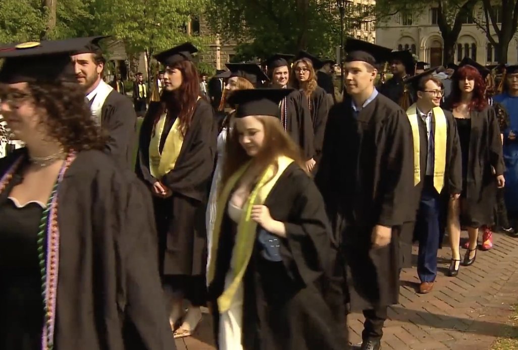 Students marching during commencement, some wearing lemon-yellow stoles with their graduation robes.
