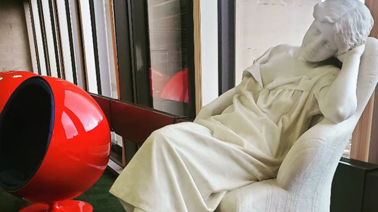 Marble statue of seated young woman, her head resting against her hand, red womb chairs in back.