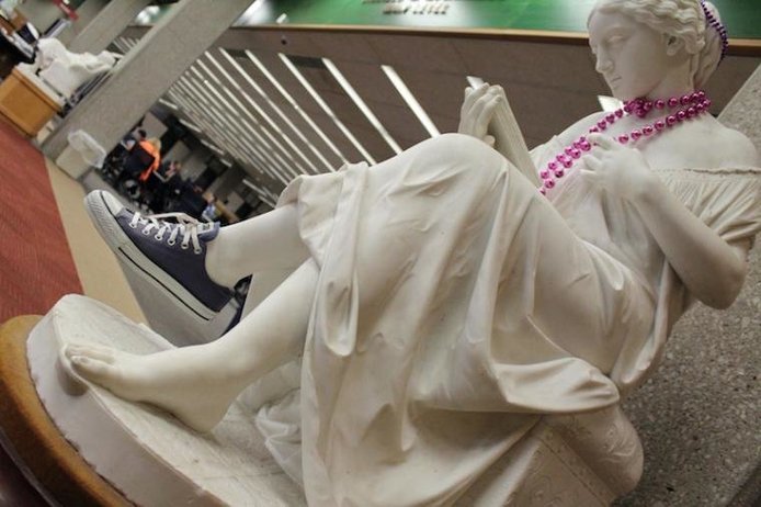 Marble sculpture of girl reading from a book with pink beads and a sneaker added to her.