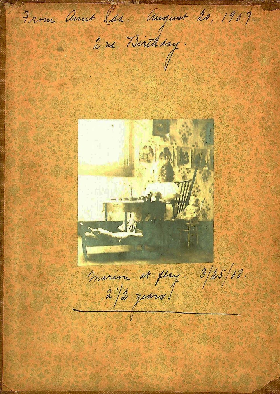 Child seated in room, sepia tone photo, pasted onto brown and green background paper with inscription.